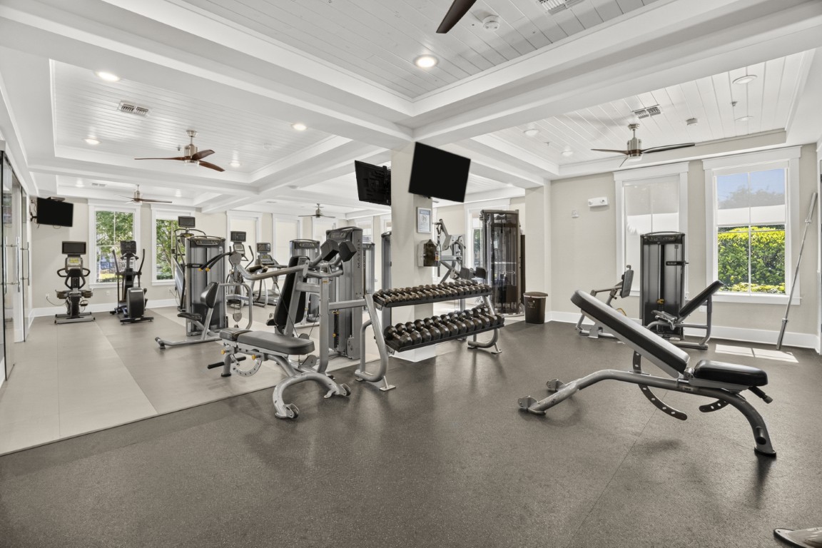 Novus Modern gym with various exercise machines, free weights, benches, mirrors, TV screens, ceiling fans, and large windows letting in sunlight.