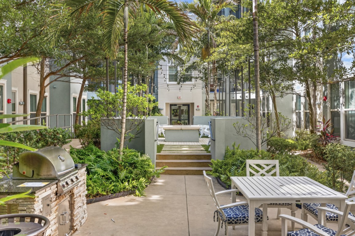 Novus Outdoor courtyard with a grill, white patio furniture, lush green plants, and steps leading to a building entrance.