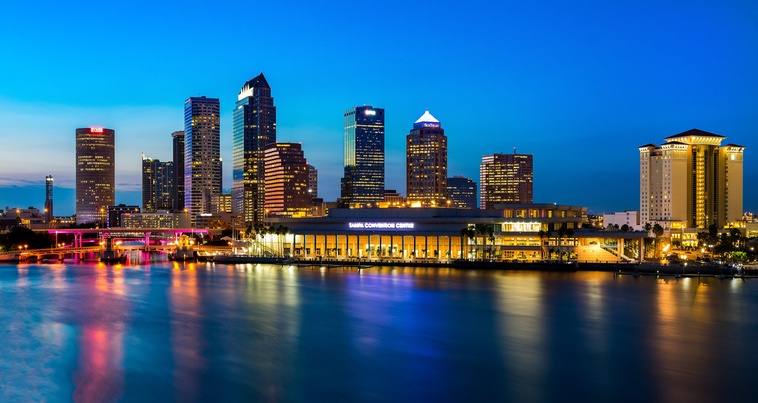 Novus City skyline at dusk with tall buildings, a convention center, and colorful reflections on calm water.