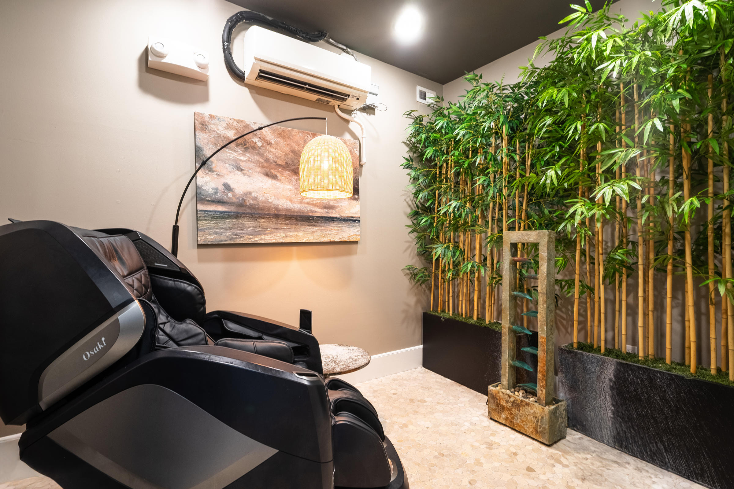 A black massage chair in a serene room with bamboo plants, a water feature, and a hanging lamp.