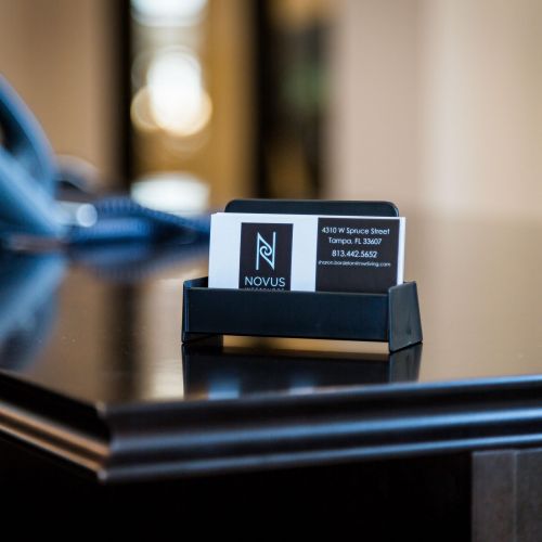 A business card in a black holder sits on a dark desk near a phone in an office setting.