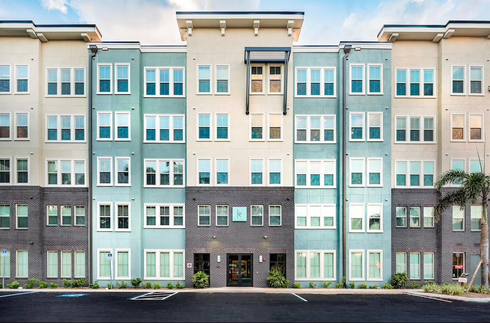 Modern four-story apartment building with many windows and a small palm tree in front.