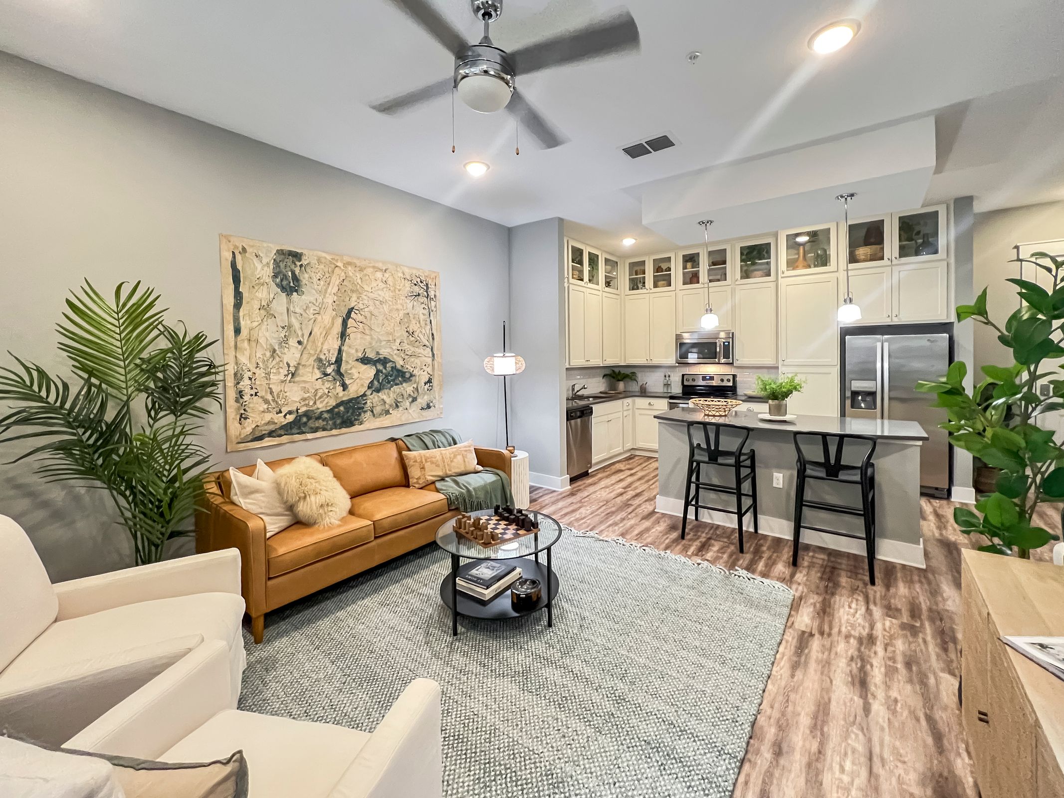Novus Modern open-concept living room and kitchen, with tan sofa, armchairs, island seating, and green plants.