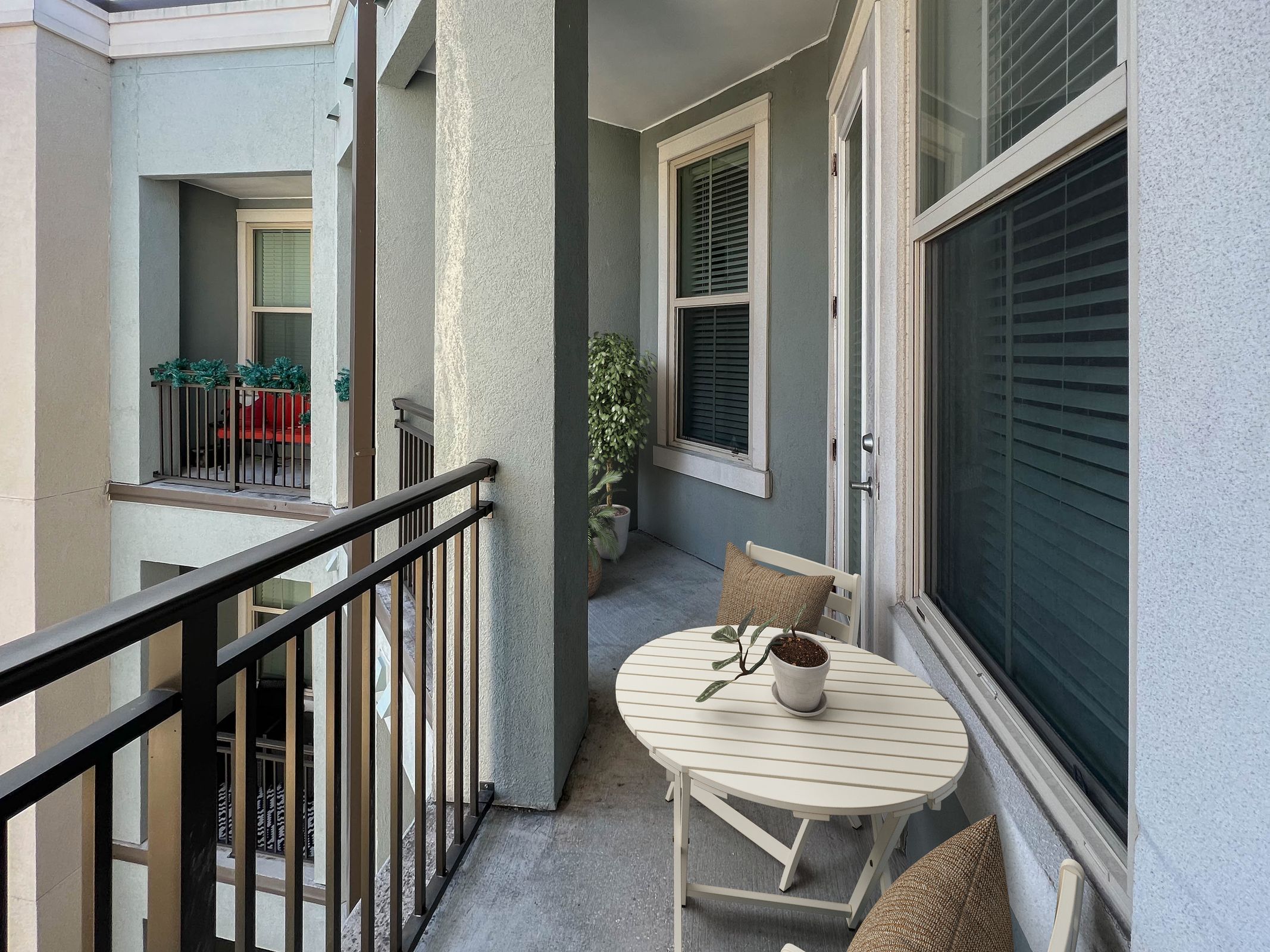 Novus A small balcony with a round white table, two chairs, a potted plant, railing, and adjacent windows.